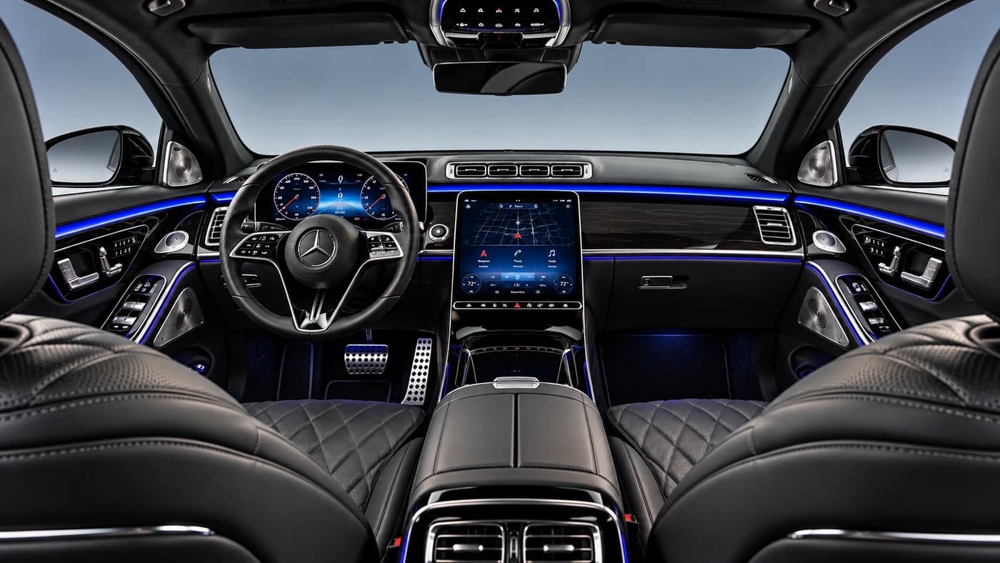 Driver's perspective of the Mercedes Benz S-Class interior showing black leather seats, blue lighting, a central screen, and wood trim.