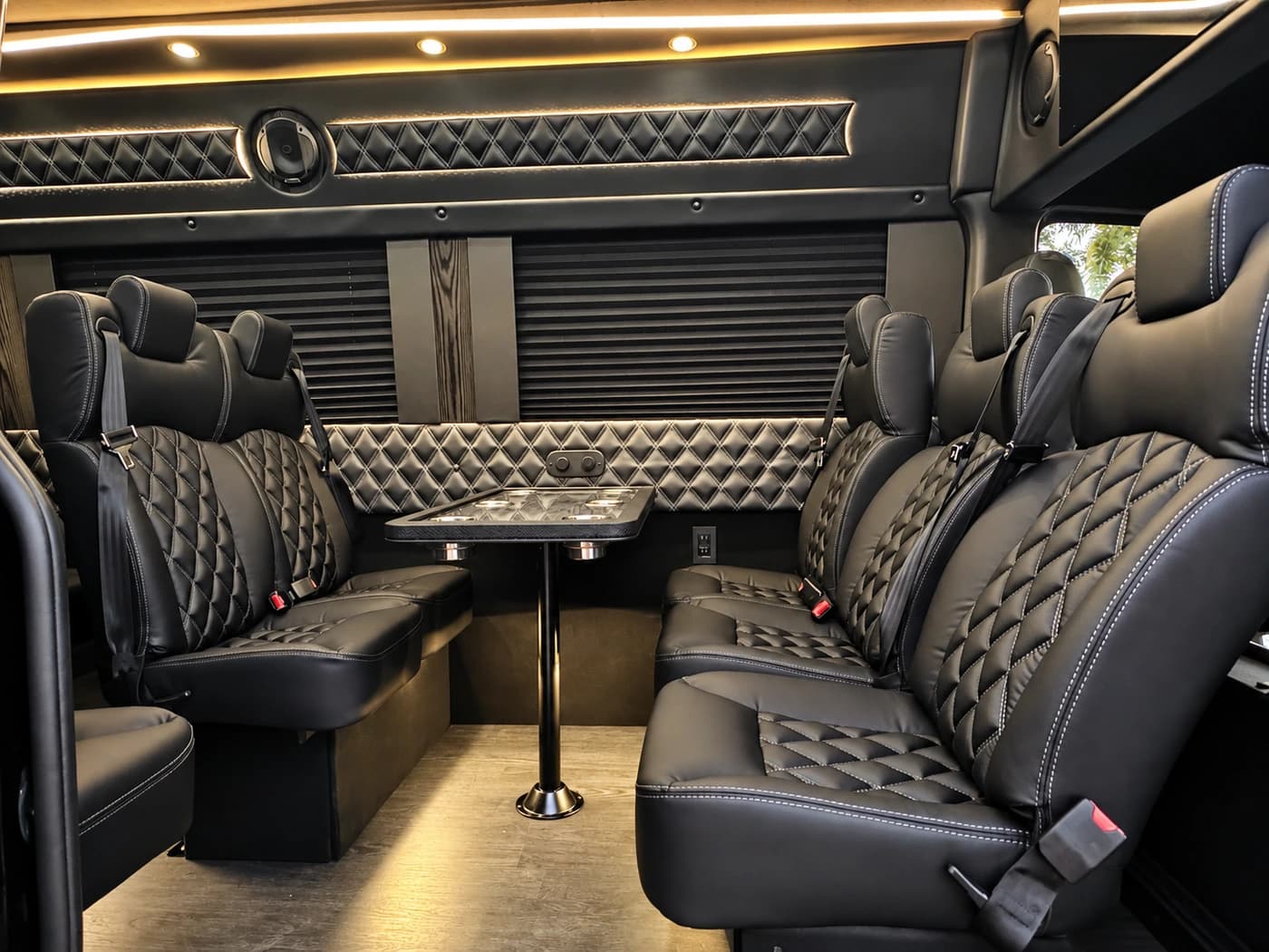 The Mercedes-Benz Executive Sprinter interior displays black diamond-quilted leather seating, a central table, and ceiling lighting.