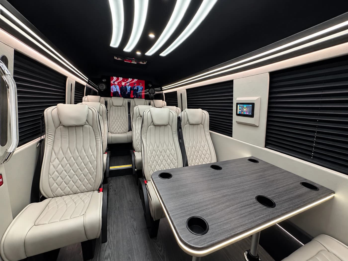 The Mercedes-Benz Executive Sprinter shows white leather seats, a dark table, black panels, curved ceiling lights, and screens.
